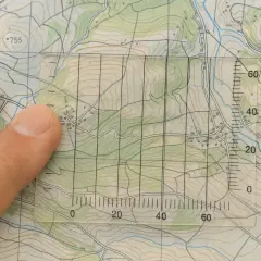 Finger überlagert eine Karte, während ein transparentes Lineal mit Gitternetz und Skalen zum **Karten lesen** verwendet wird.