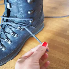 Eine Hand mit roten Nägeln bindet die Schnürsenkel eines robusten, dunklen Wanderschuhs. Bereit für das nächste Abenteuer.