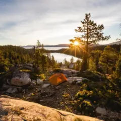 Ein orangefarbenes Zelt steht auf einem felsigen Hang mit Blick auf einen See, umgeben von Bäumen. Die Sonne bricht durch einen Baum und erzeugt einen Sterneneffekt. Ein Traum für jeden, der **wildcampen Europa** liebt.