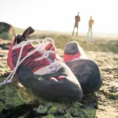 Boulderschuhe ruhen auf moosbewachsenem Fels. Im Hintergrund stehen zwei Kletterer. Zeit für eine Wäsche nach dem Abenteuer.