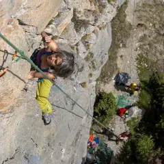 Frau klettert in Briançon, konzentriert sich auf den nächsten Griff. Unten warten Kletterer und Ausrüstung.