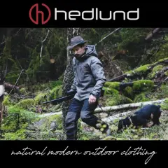 Ein Mann in Lodenkleidung und Hut geht mit einem Hund und einem Gewehr durch einen moosbewachsenen Wald.