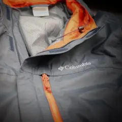 Eine graue Columbia Hardshelljacke mit orangefarbenem Reißverschluss und Kragen. Perfekt für jedes Wetter.