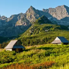 Zwei Holzhütten in einer grünen Bergwiese, im Hintergrund die majestätischen Gipfel der Hohen Tatra.