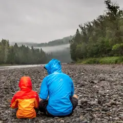 Zwei Personen in bunten Regenjacken sitzen am steinigen Ufer eines Flusses. Die richtige Gore-Tex-Pflege hält sie trocken.