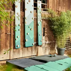 DIY-Kletterwand outdoor: Grüne Bretter mit Holzgriffen an Holzwand, daneben eine Schaukel und Klettermatten.