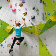Eine Frau klettert an einer bunten Boulderwand. Sie greift nach Griffen, um sich hochzuziehen. Das ist, was ist bouldern: Klettern ohne Seil in Absprunghöhe.