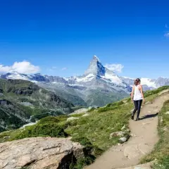 Frau wandert auf einem Pfad mit Blick auf das majestätische Matterhorn. Die Berglandschaft ist grün und felsig, der Himmel strahlend blau.