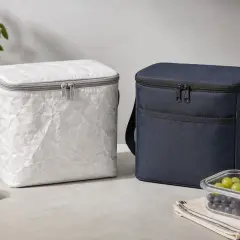 Zwei Kühltaschen, eine in weißem, zerknittertem rPET-Material, die andere in dunkelblauem Stoff. Daneben eine Flasche, Zitrone und eine Obstschale.
