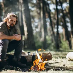 Mann sitzt am Lagerfeuer im Wald, hält eine Tasse. Das ist Bushcraft: Naturverbundenheit und Selbstversorgung.