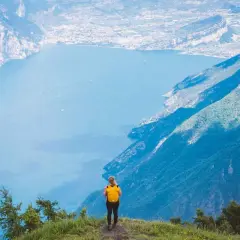 Frau mit gelbem Rucksack steht auf einem Berg und blickt auf einen See. Ein traumhafter Ausblick auf die italienischen Fernwanderwege.
