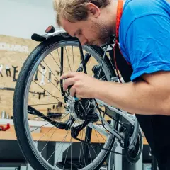 Ein Mann mit Bart im blauen T-Shirt und schwarzer Schürze arbeitet konzentriert am Fahrrad. Er wechselt das Fahrrad Schaltwerk.