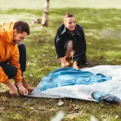 Mann und Frau bauen ein Zelt auf. Sie legen die Zeltplane aus und befestigen sie im Gras.