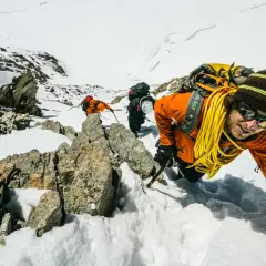 Bergsteiger kämpfen sich durch den Schnee in den Berner Alpen. Ein Mann in Orange lächelt in die Kamera, während er sich an einem Felsen festhält.