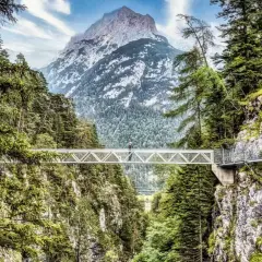 Ein Wanderer überquert eine Hängebrücke imalat, umgeben von grünen Wäldern und mit einem majestätischen Berg im Hintergrund.