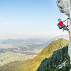 Ein Mann klettert gesichert an einem Stahlseil einen Felsvorsprung entlang. Der Klettersteig in Deutschland bietet eine atemberaubende Aussicht auf das Tal.