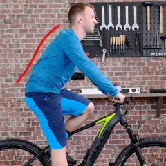 Mann auf einem Fahrrad in aufrechter Sitzposition, der Rücken ist leicht gekrümmt. Werkzeugwand im Hintergrund.