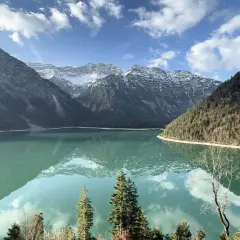 Spiegelglatter See am Plansee Panoramaweg, umrahmt von bewaldeten Bergen und schneebedeckten Gipfeln. Ein perfekter Tag für eine Wanderung.
