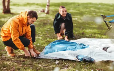 Mann und Frau bauen ein Zelt auf. Sie legen die Zeltplane aus und befestigen sie im Gras.