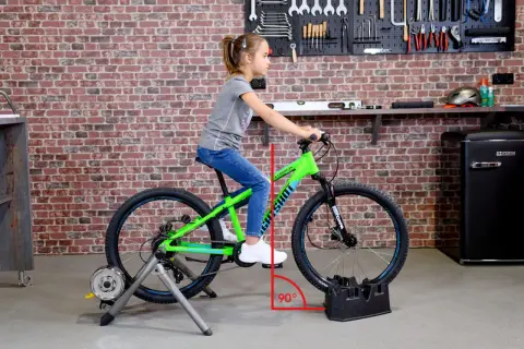 Ein Mädchen sitzt auf einem grünen Kinderfahrrad, das auf einem Rollentrainer steht. Eine rote Linie zeigt den 90-Grad-Winkel für das richtige Einstellen des Kinderfahrrads.