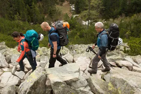 Drei Wanderer mit Rucksäcken und Helmen auf felsigem Untergrund. Die Ausrüstung auf den Rücken deutet auf eine Klettersteig-Packliste hin.