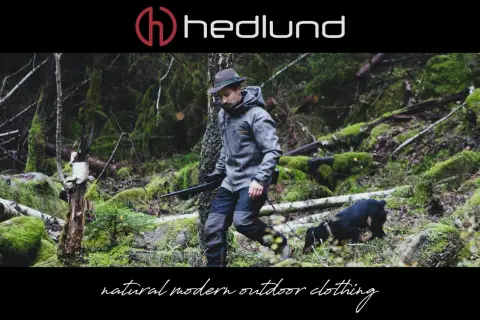 Ein Mann in Lodenkleidung und Hut geht mit einem Hund und einem Gewehr durch einen moosbewachsenen Wald.