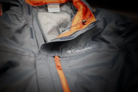 Eine graue Columbia Hardshelljacke mit orangefarbenem Reißverschluss und Kragen. Perfekt für jedes Wetter.