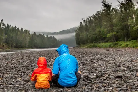 Zwei Personen in bunten Regenjacken sitzen am steinigen Ufer eines Flusses. Die richtige Gore-Tex-Pflege hält sie trocken.
