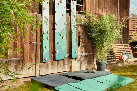 DIY-Kletterwand outdoor: Grüne Bretter mit Holzgriffen an Holzwand, daneben eine Schaukel und Klettermatten.