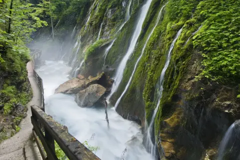 Ein Pfad führt entlang einer tosenden Klamm in Bayern, wo Wasserfälle über moosbewachsene Felsen stürzen.