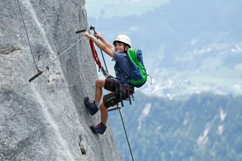 Ein Junge mit Rucksack und Helm meistert einen Klettersteig. Ein tolles Abenteuer für klettersteig kinder!