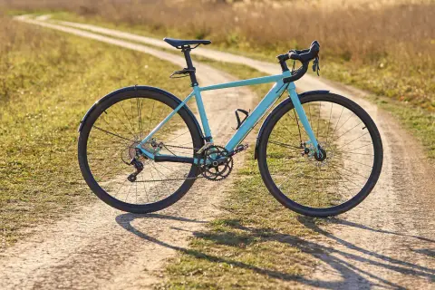 Ein hellblaues Fahrrad mit Schutzblechen steht auf einem Feldweg. Der Unterschied zwischen Cyclocross und Gravel wird hier deutlich: Dieses Rad ist bereit für Abenteuer auf Schotterwegen.