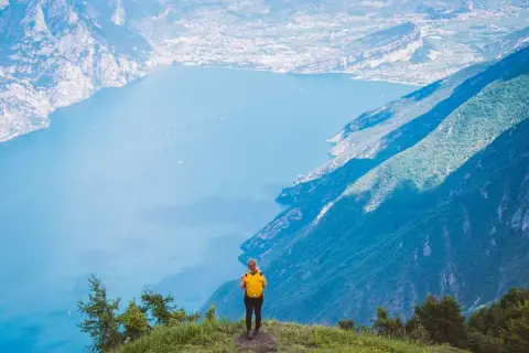Frau mit gelbem Rucksack steht auf einem Berg und blickt auf einen See. Ein traumhafter Ausblick auf die italienischen Fernwanderwege.