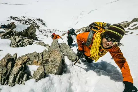 Bergsteiger kämpfen sich durch den Schnee in den Berner Alpen. Ein Mann in Orange lächelt in die Kamera, während er sich an einem Felsen festhält.