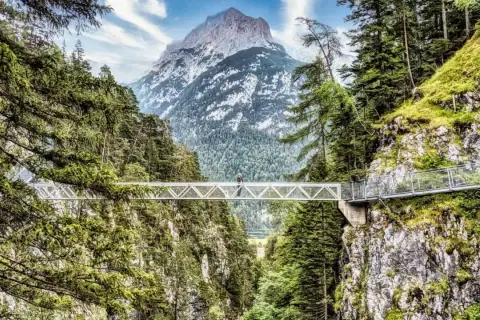 Ein Wanderer überquert eine Hängebrücke imalat, umgeben von grünen Wäldern und mit einem majestätischen Berg im Hintergrund.
