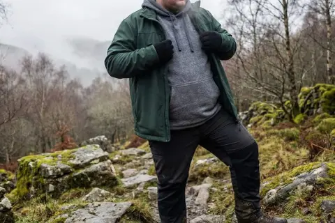 Mann in grüner Jacke und grauer Kapuzenjacke wandert durch felsiges Gelände. Die Jacke ist softshell wasserdicht und schützt vor dem Wetter.
