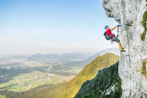 Ein Mann klettert gesichert an einem Stahlseil einen Felsvorsprung entlang. Der Klettersteig in Deutschland bietet eine atemberaubende Aussicht auf das Tal.