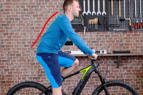 Mann auf einem Fahrrad in aufrechter Sitzposition, der Rücken ist leicht gekrümmt. Werkzeugwand im Hintergrund.