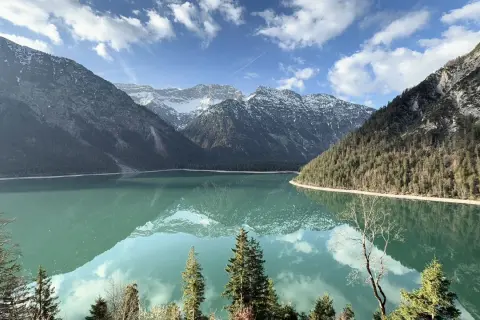 Spiegelglatter See am Plansee Panoramaweg, umrahmt von bewaldeten Bergen und schneebedeckten Gipfeln. Ein perfekter Tag für eine Wanderung.