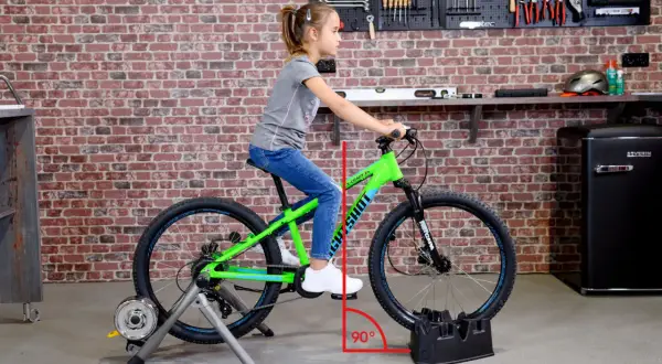 Ein Mädchen sitzt auf einem grünen Kinderfahrrad, das auf einem Rollentrainer steht. Eine rote Linie zeigt den 90-Grad-Winkel für das richtige Einstellen des Kinderfahrrads.