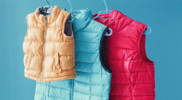Drei Steppwesten in Orange, Türkis und Pink hängen an Kleiderbügeln. So waschen Sie Ihre daunenjacke waschen ohne trockner.
