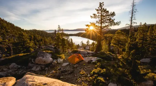 Ein orangefarbenes Zelt steht auf einem felsigen Hang mit Blick auf einen See, umgeben von Bäumen. Die Sonne bricht durch einen Baum und erzeugt einen Sterneneffekt. Ein Traum für jeden, der **wildcampen Europa** liebt.