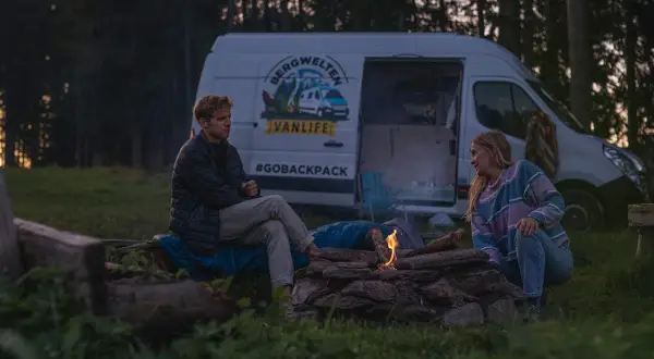 Ein Paar genießt das Lagerfeuer beim wildcampen Bayern. Ihr Van steht im Hintergrund, bereit für das nächste Abenteuer.