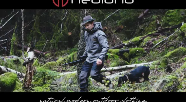 Ein Mann in Lodenkleidung und Hut geht mit einem Hund und einem Gewehr durch einen moosbewachsenen Wald.