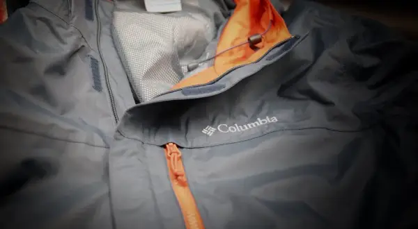 Eine graue Columbia Hardshelljacke mit orangefarbenem Reißverschluss und Kragen. Perfekt für jedes Wetter.