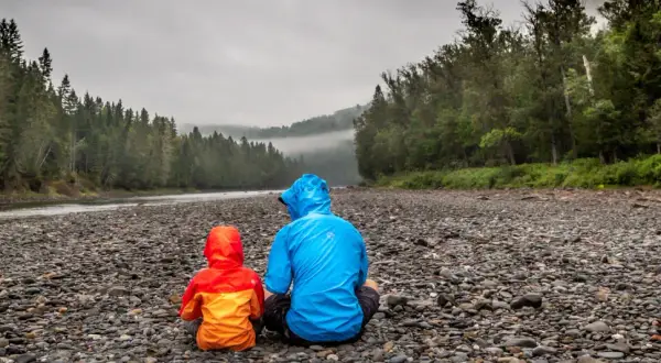 Zwei Personen in bunten Regenjacken sitzen am steinigen Ufer eines Flusses. Die richtige Gore-Tex-Pflege hält sie trocken.