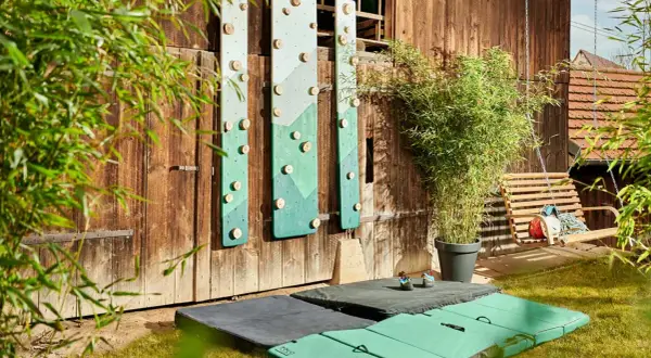 DIY-Kletterwand outdoor: Grüne Bretter mit Holzgriffen an Holzwand, daneben eine Schaukel und Klettermatten.
