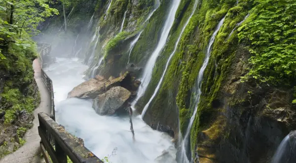 Ein Pfad führt entlang einer tosenden Klamm in Bayern, wo Wasserfälle über moosbewachsene Felsen stürzen.