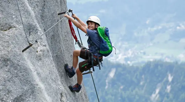 Ein Junge mit Rucksack und Helm meistert einen Klettersteig. Ein tolles Abenteuer für klettersteig kinder!