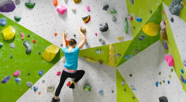Eine Frau klettert an einer bunten Boulderwand. Sie greift nach Griffen, um sich hochzuziehen. Das ist, was ist bouldern: Klettern ohne Seil in Absprunghöhe.